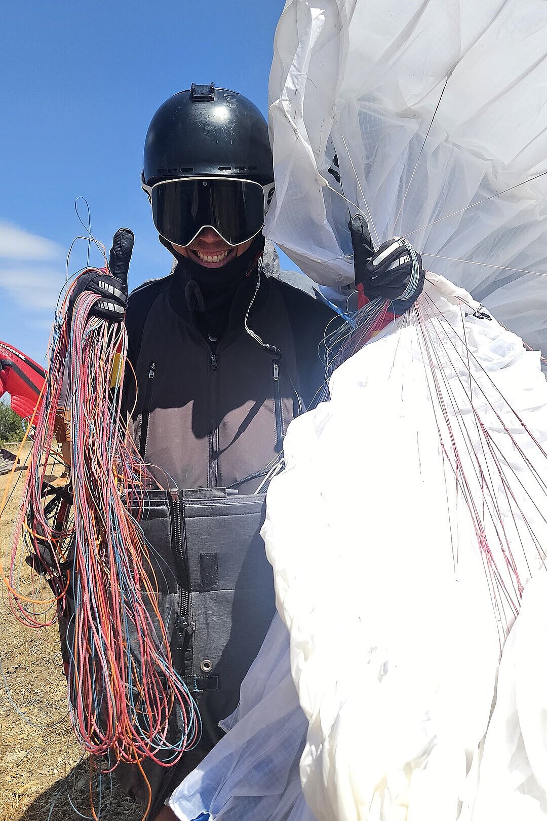 Person mit schwarzem Helm und Skibrille zeigt den Daumen hoch und hält Leinen neben einem weißen Schirm.