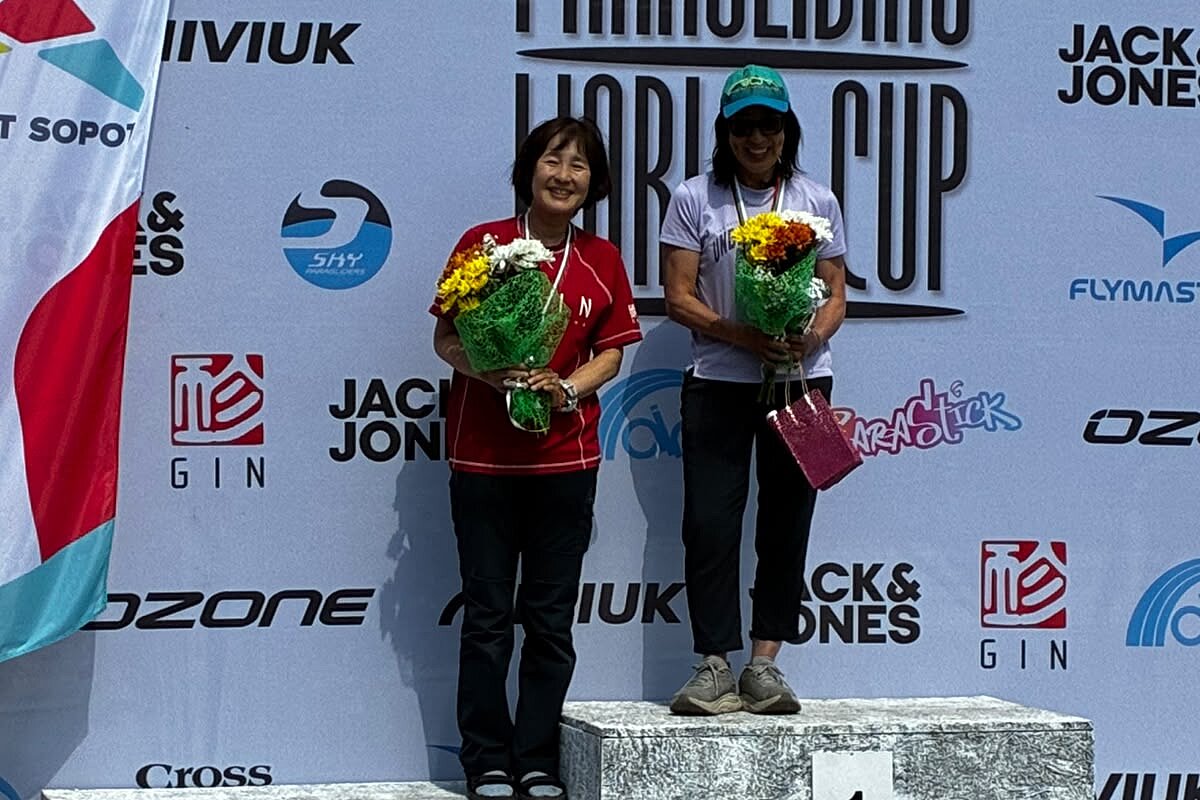 Zwei Frauen auf dem Siegerpodest mit Blumensträußen vor einer Paragliding-Weltcup-Wand.