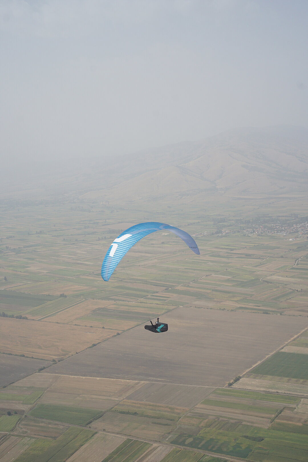 Ein einzelner blauer Gleitschirm fliegt über Felder und ein kleines Dorf im Dunst.