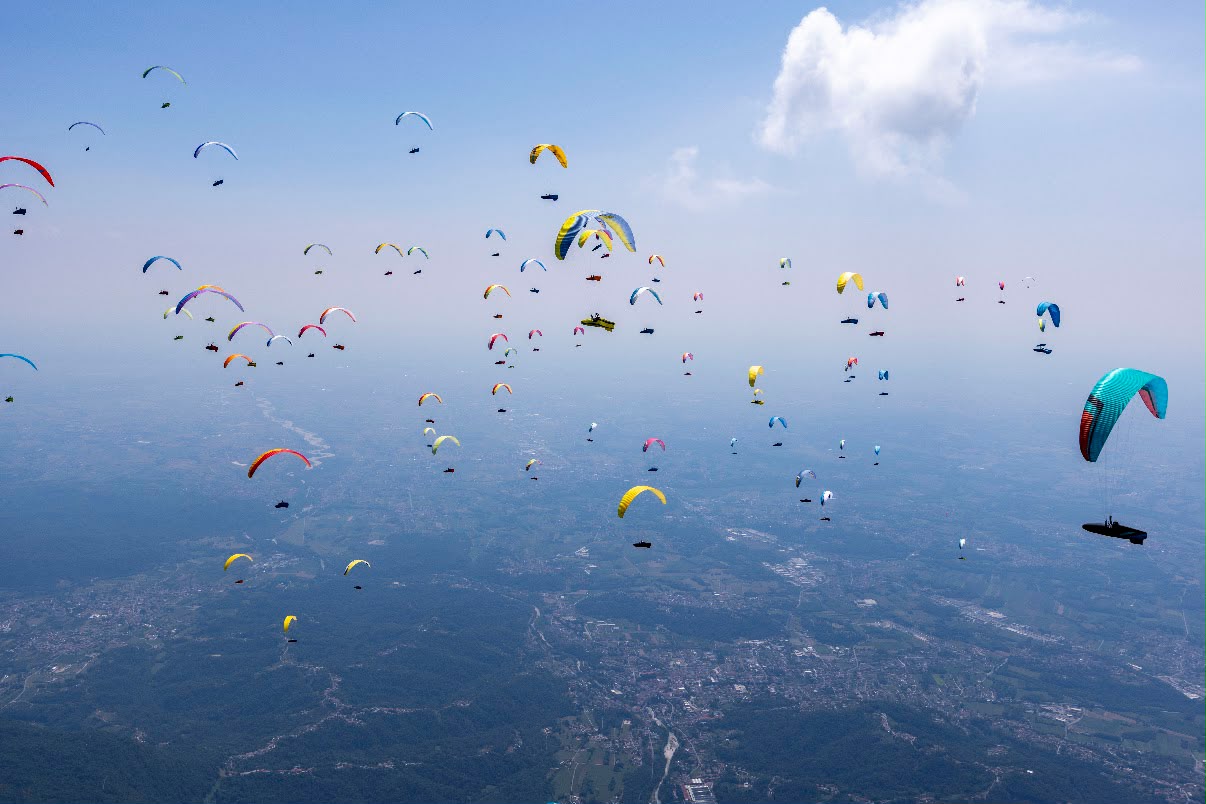 Viele Gleitschirme fliegen über einer weiten Landschaft, rechts ein großer türkisfarbener Schirm nah im Bild.