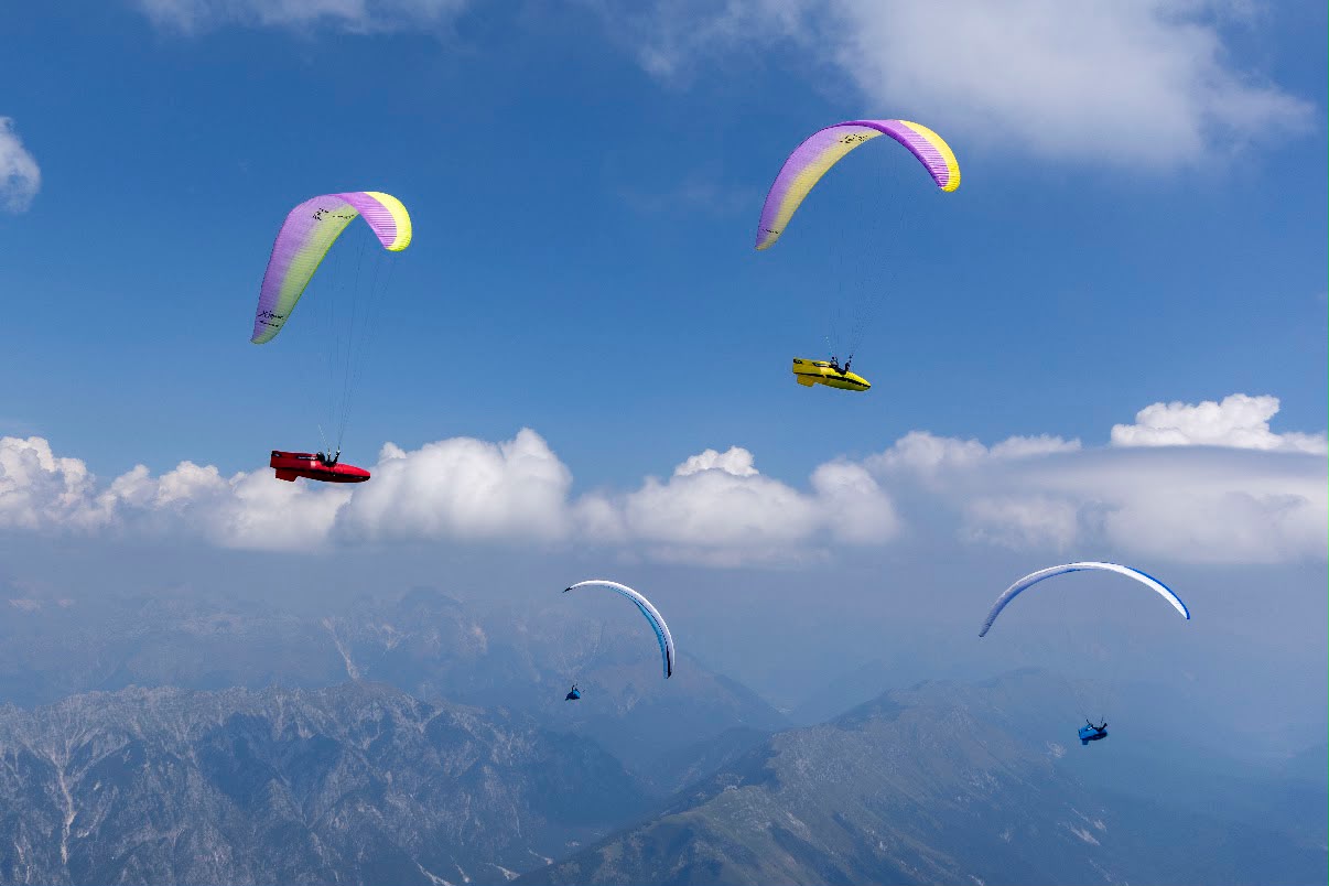 Mehrere Gleitschirme schweben vor blauem Himmel über Bergkämmen, darunter ein roter und ein gelber Schirm.